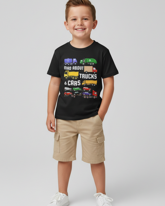Young boy model wearing a black trucks and cars graphic T-shirt with beige cargo shorts and white trainers, standing against a clean white studio background.