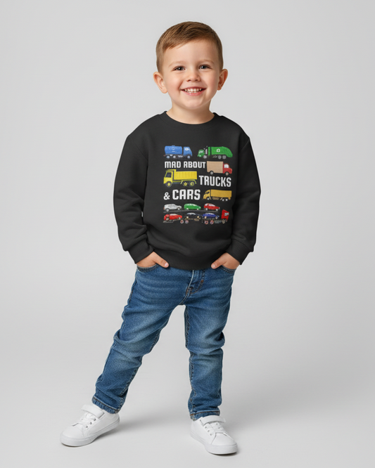Smiling young boy wearing black “Mad About Trucks & Cars” kids sweatshirt with colourful vehicle graphic, styled with blue jeans and white trainers on a studio background