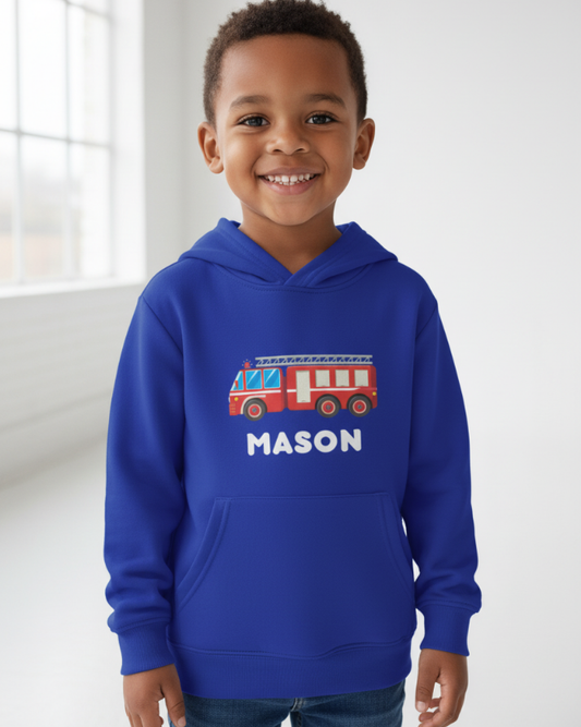 Young boy wearing royal blue personalised fire truck hoodie with custom name “Mason”, smiling in bright studio setting, graphic fully visible on front.