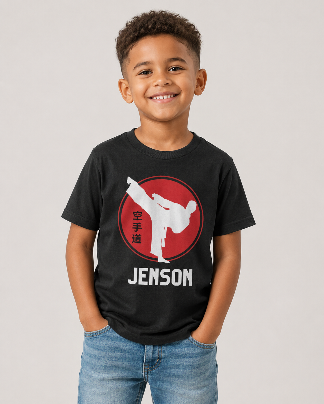 A young boy smiling and wearing a black graphic t-shirt with a red circular martial arts/karate design and the name 'JENSON' printed in white, paired with blue jeans, against a light grey background.