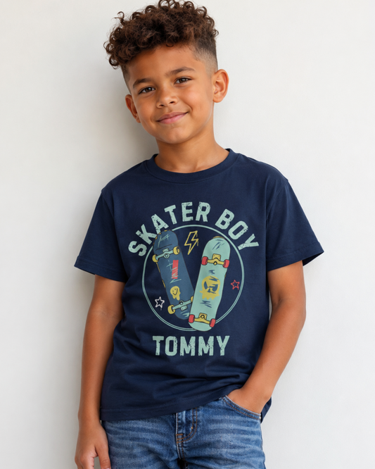 A young boy with curly hair wearing a navy blue graphic t-shirt that reads 'SKATER BOY' with custom name 'TOMMY' with a skateboard design, paired with blue jeans, standing against a plain white wall.