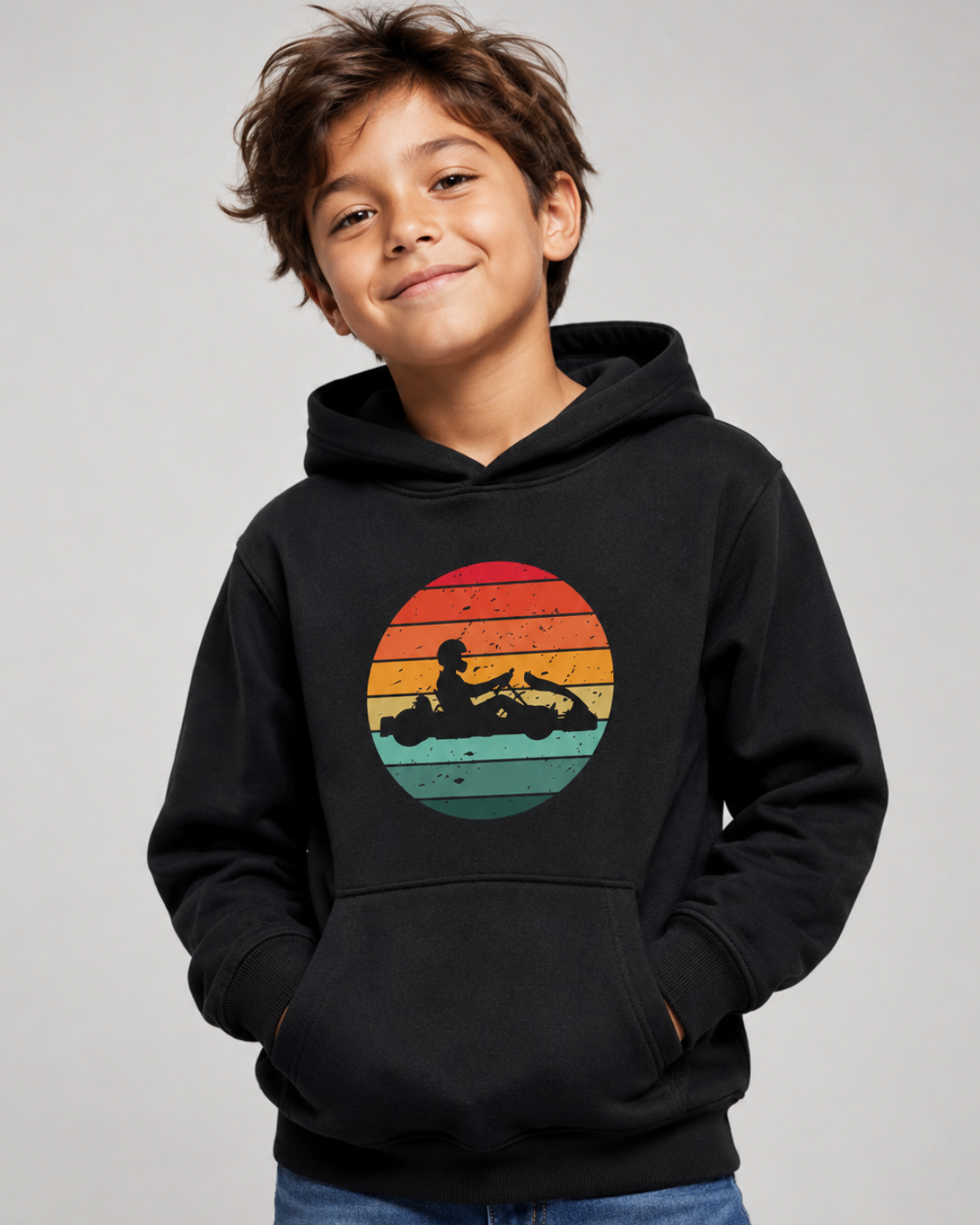A young boy smiling while wearing a black pullover hoodie with a retro sunset graphic on the chest with Go-kart driver silhouette, paired with blue jeans, photographed against a neutral grey studio background.