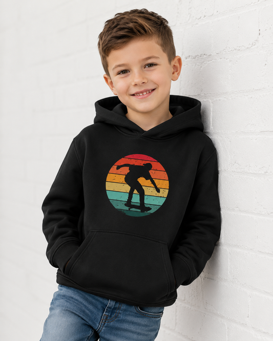 A young boy smiling and leaning against a white brick wall, wearing a black hoodie with a retro sunset graphic of a skateboarder on the chest, paired with blue jeans.