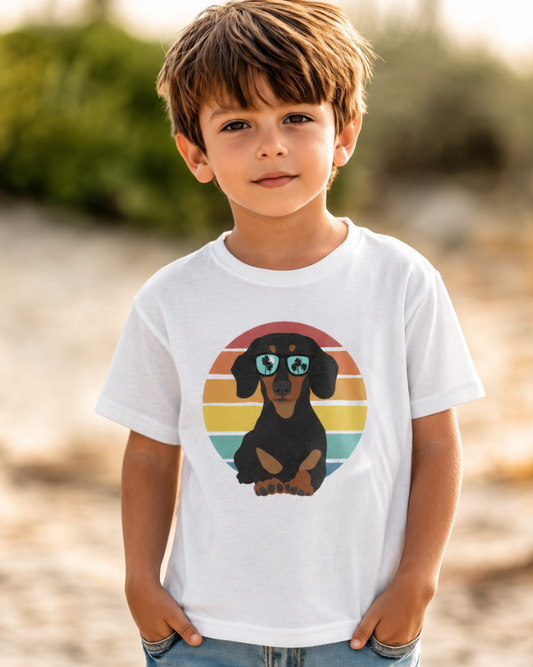 Boy at the beach wearing a kids short sleeved white t-shirt with a retro dachshund in sunglasses graphic.