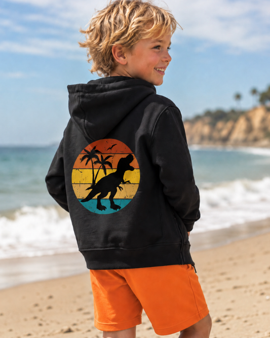Boy on the beach wearing a black zip hoodie with retro sunset T-Rex back print and orange shorts.