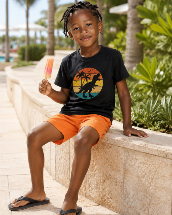 Boys summer dinosaur t-shirt in black, worn with orange shorts in a sunny resort setting