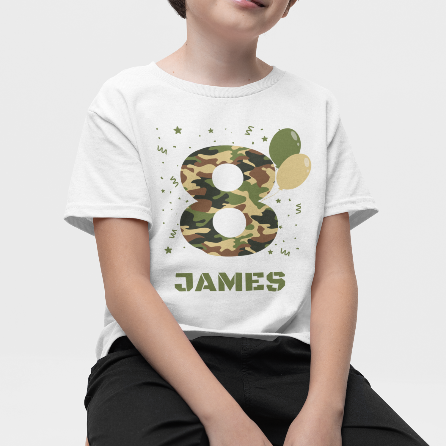 Boy sitting on a stool wearing a personalised camouflage number 8 birthday t-shirt with name Jamie, white kids birthday tee with green camo design