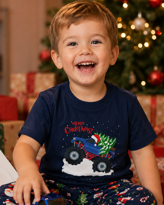 Little boy wearing a Christmas-themed t-shirt with a blue and red monster truck and tree design, sitting in front of a decorated Christmas tree.