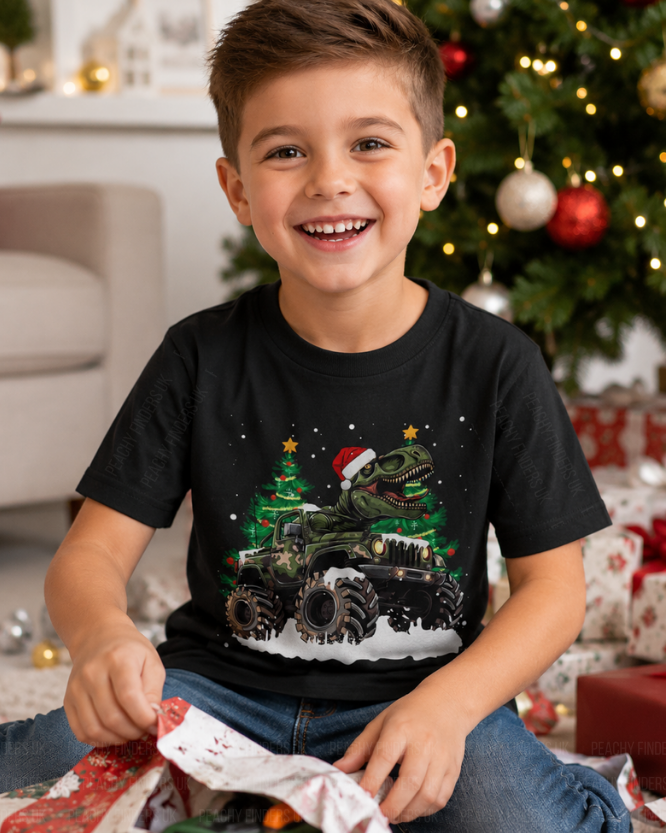 Young boy wearing a black t-shirt with a Christmas-themed graphic of a t-rex riding a monster truck, sitting in front of a decorated Christmas tree.