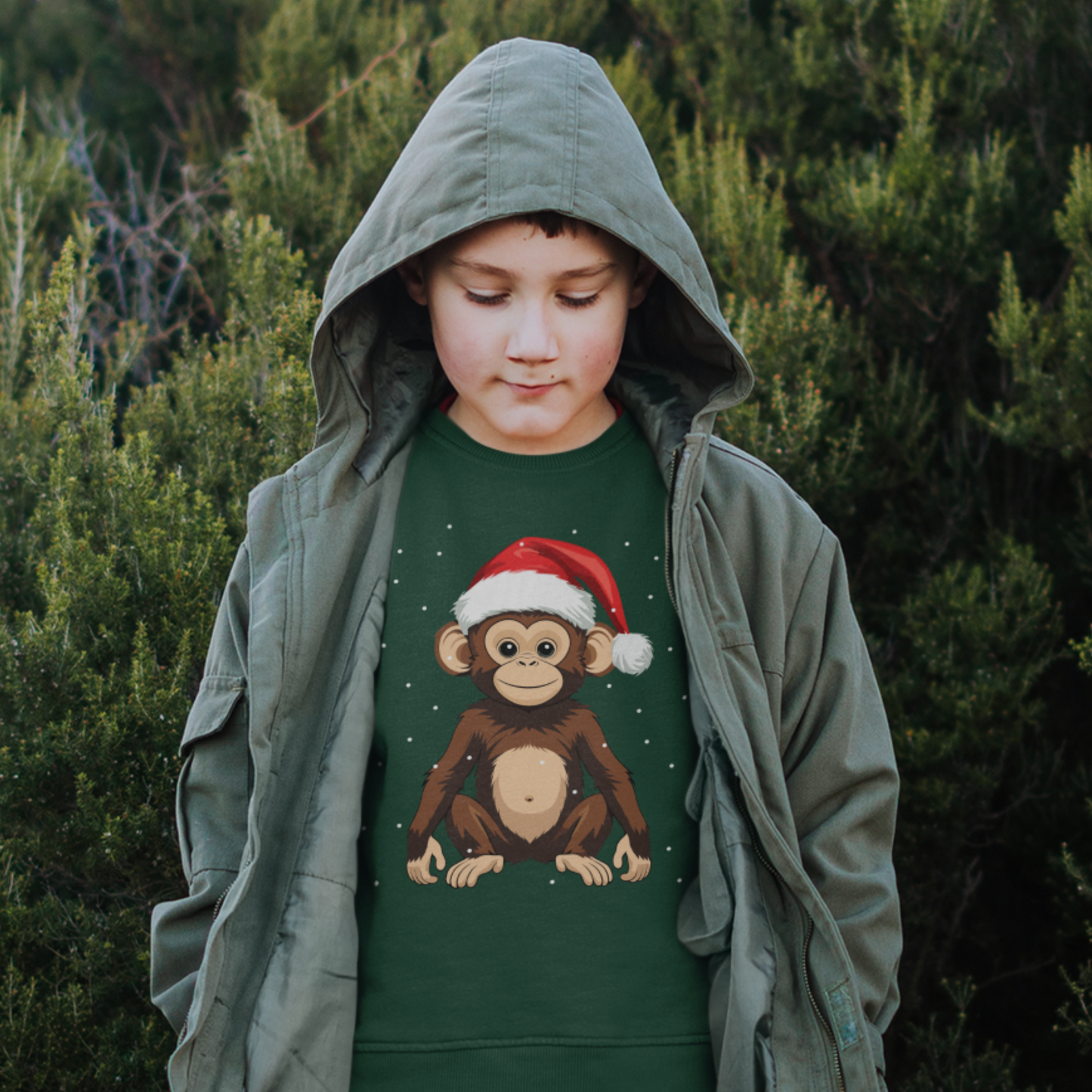 A young boy wearing a green jumper with a Christmas-themed monkey design, styled under a green jacket with hood up in a forest setting