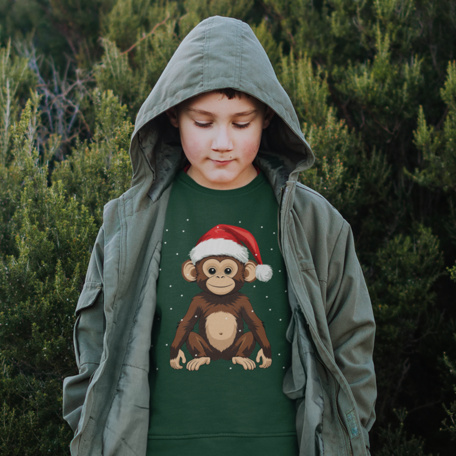 A young boy wearing a green jumper with a Christmas-themed monkey design, styled under a green jacket with hood up in a forest setting