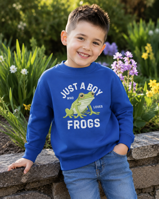 A young boy smiling outdoors in a garden, wearing a bright royal blue crewneck sweatshirt with a graphic that reads 'JUST A BOY WHO LOVES FROGS' above and below a green frog illustration, paired with blue jeans.