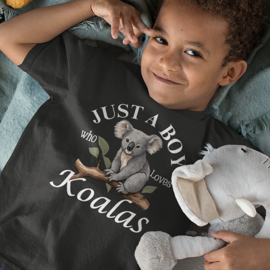 Young boy wearing a black T-shirt featuring a printed design of a cute koala sitting on a tree branch with the words “Just A Boy Who Loves Koalas” in white text.