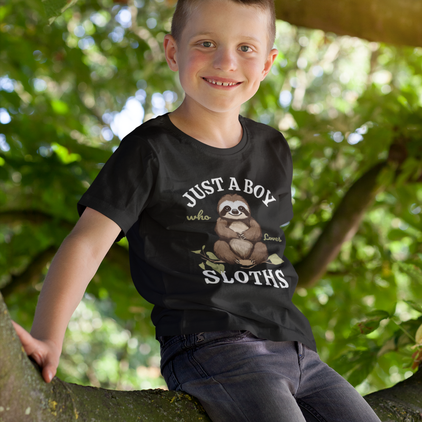 Boy sitting in a tree wearing a black “Just A Boy Who Loves Sloths” T-shirt with a cute brown sloth sitting on a leafy branch printed on the front, smiling at the camera.