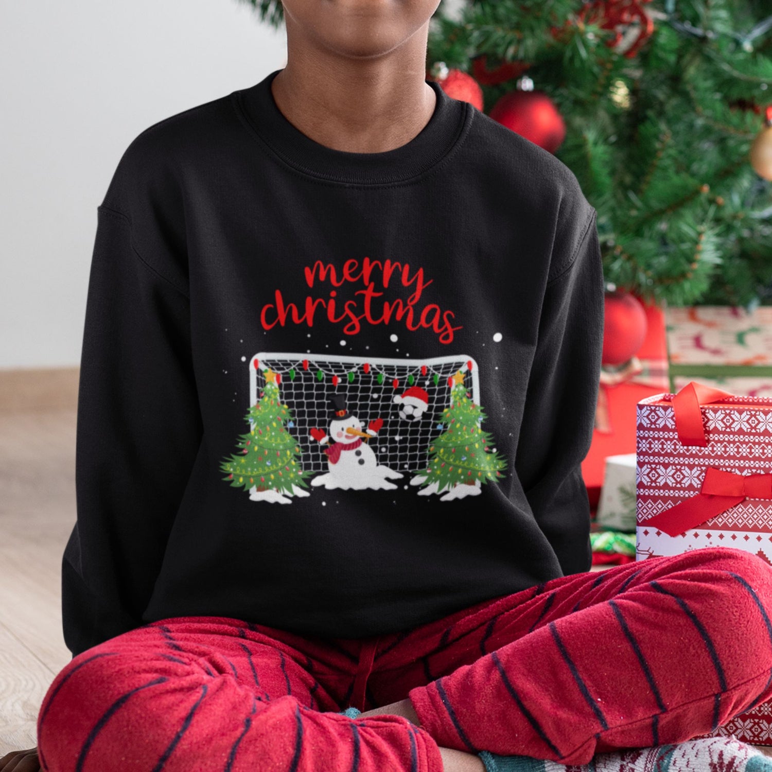 Boy wearing black Christmas sweatshirt with snowman goalkeeper and football goal design, festive Christmas trees, and “Merry Christmas” text.