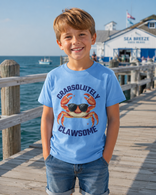 A young boy smiling on a wooden pier wearing a light blue t-shirt with a graphic of a crab wearing sunglasses and the text 'CRABSOLUTELY CLAWSOME'.