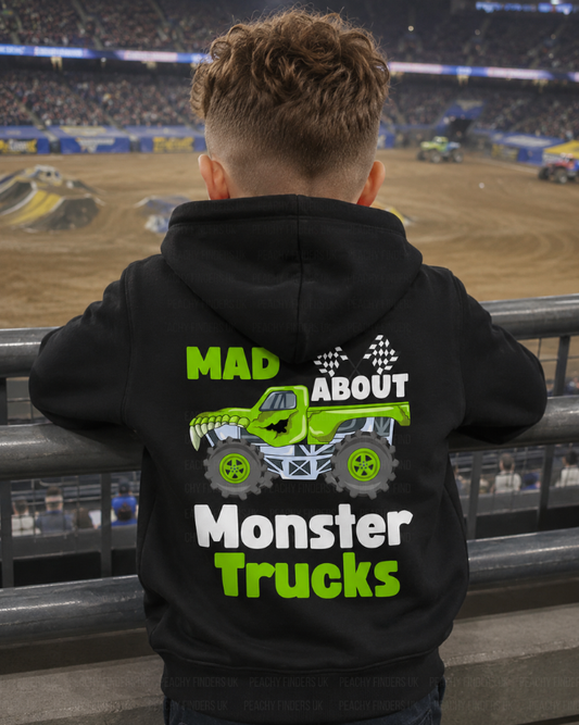 Young boy wearing a black kids hoodie with bold “Mad About Monster Trucks” back print, leaning over arena railings at a monster truck show.