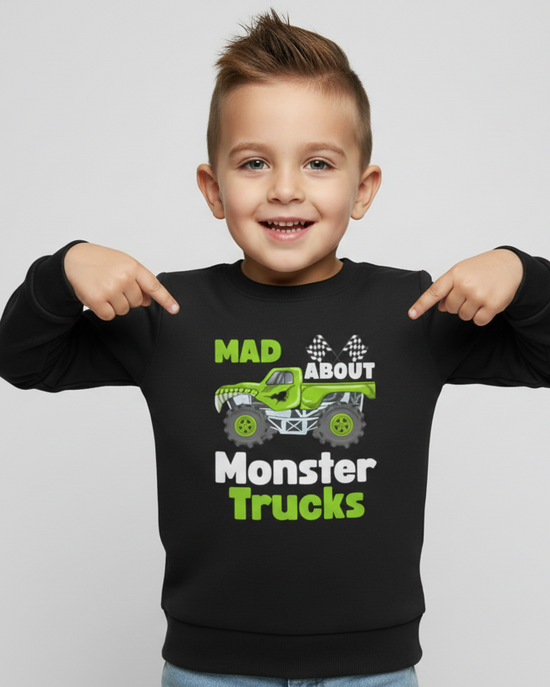 Young boy wearing a black kids sweatshirt with green monster truck graphic and the slogan “Mad About Monster Trucks”, pointing at the design, studio background