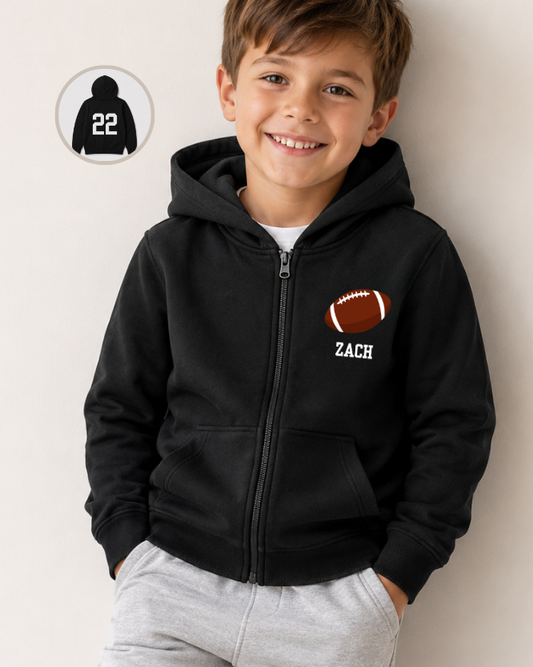 A smiling young boy wearing a black zip-up hoodie with a custom football graphic and the name 'ZACH' printed on the chest. The image features a light gray background and includes a circular inset graphic in the top-left corner showing the back of the hoodie with the number '22'.