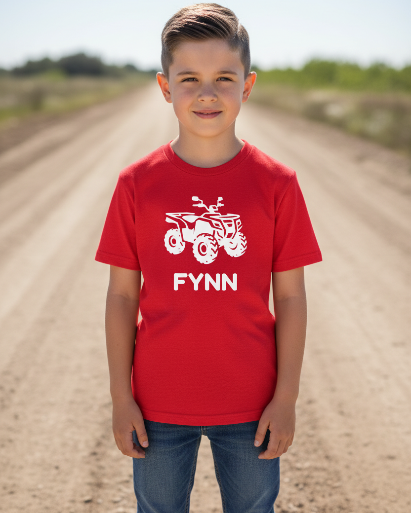 Boy outdoors on a dirt track wearing a red personalised quad bike kids t-shirt with white ATV graphic and the name “Fynn”.