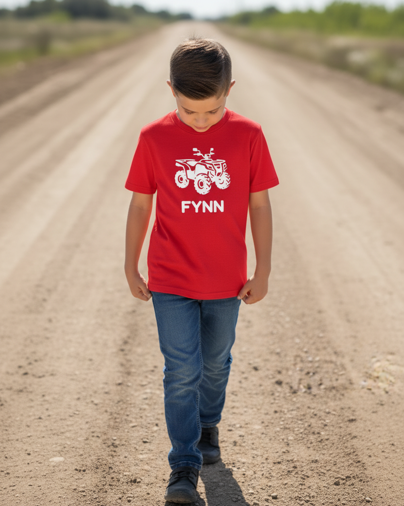 Boy walking outdoors on a dirt track wearing a red custom name quad bike t-shirt with white ATV silhouette and the name “Fynn”.