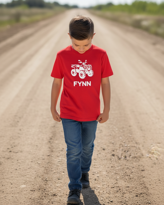Boy walking outdoors on a dirt track wearing a red custom name quad bike t-shirt with white ATV silhouette and the name “Fynn”.