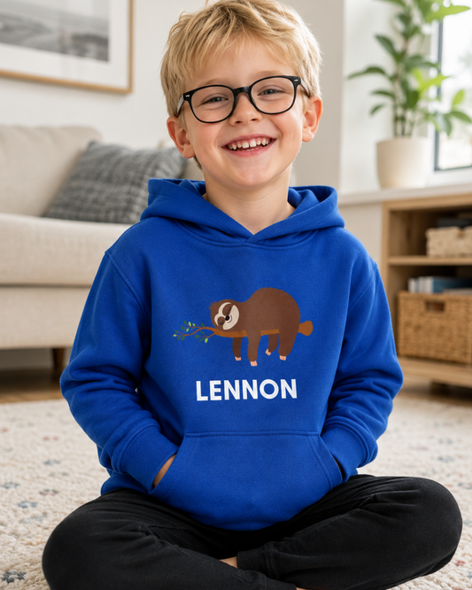A young boy with glasses sits cross-legged on a carpeted floor in a bright living room, wearing a royal blue hoodie with a cartoon sleeping sloth graphic and the name 'LENNON' printed on the front, paired with black pants.