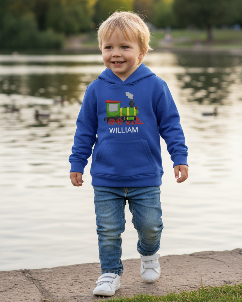 A little boy wearing a royal blue hoodie with a steam train design and name 'William' by a lake.