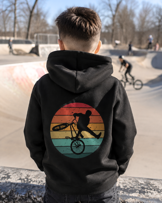 A boy viewed from behind wearing a black hoodie with a large circular retro sunset graphic on the back, featuring a BMX rider silhouette. The background shows a blurred outdoor skatepark with concrete ramps and other cyclists.