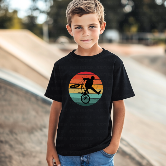 Young boy wearing a black kids t-shirt with BMX stunt rider silhouette inside a colourful retro sunset circle at a skate park