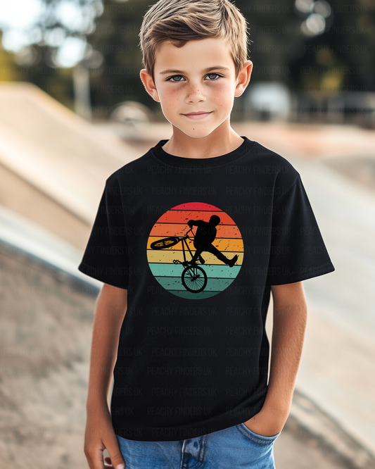 Young boy wearing a black kids t-shirt with BMX stunt rider silhouette inside a colourful retro sunset circle at a skate park
