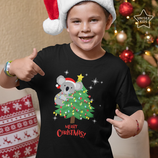 A young boy wearing a Koala Christmas T-shirt featuring a printed design of a koala wearing a Santa hat climbing a Christmas tree decorated with colourful lights and topped with a star, with the words “Merry Christmas!” below.