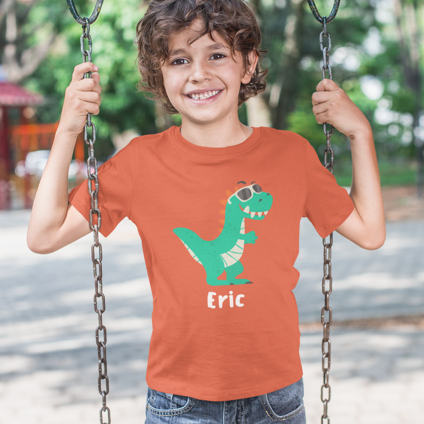 Smiling boy on a swing wearing an orange kids T-shirt with a green dinosaur in sunglasses and the personalised name Eric printed underneath.