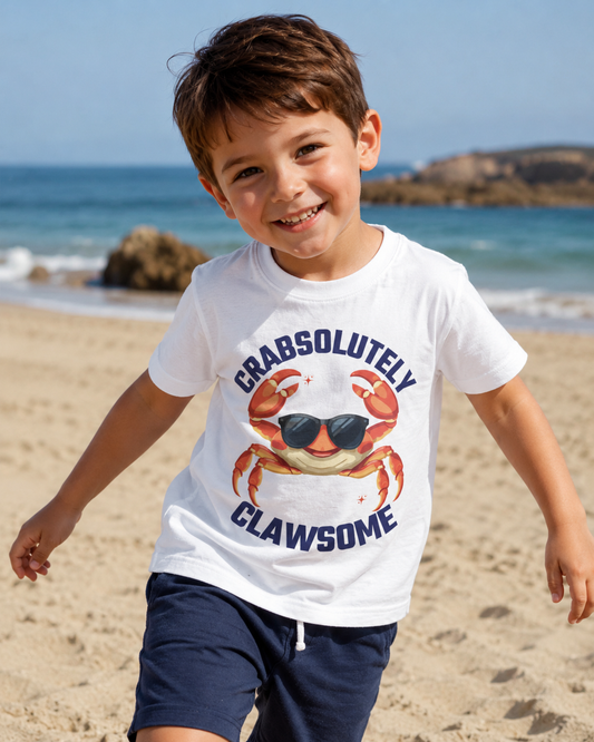 A smiling young boy running on a sandy beach wearing a white graphic t-shirt with a crab wearing sunglasses and the text 'CRABSOLUTELY CLAWSOME', paired with navy shorts.