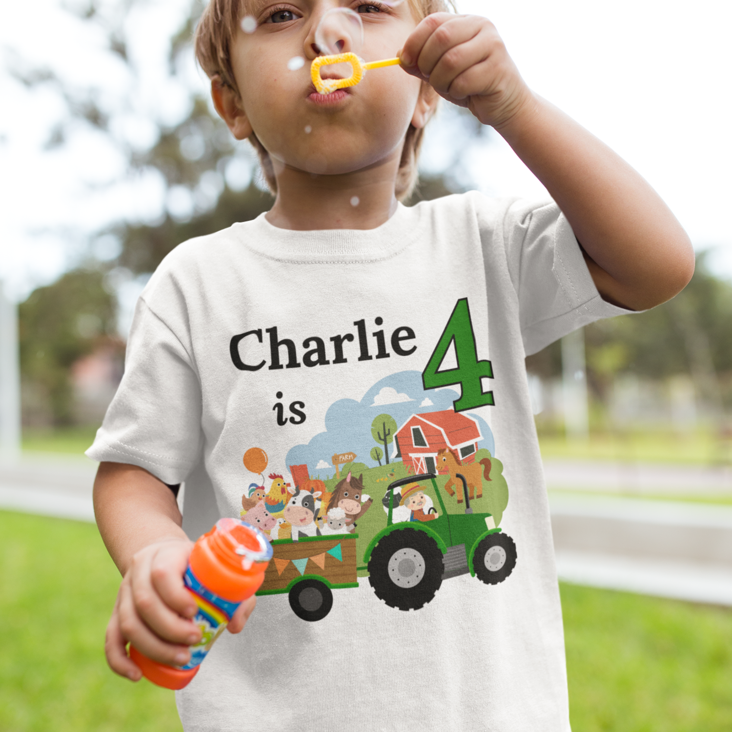 Toddler blowing bubbles while wearing a white personalised farm tractor birthday T-shirt with “Charlie is 4” and a colourful tractor and animal scene.