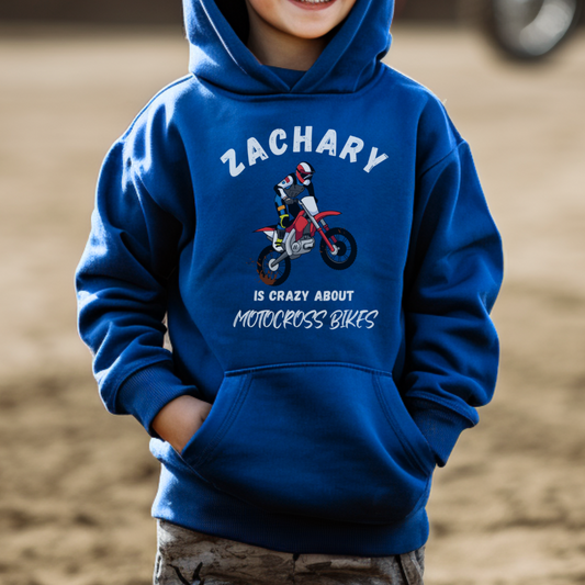 Child wearing a blue hoodie with a graphic and text about motocross bikes, standing on a dirt track.