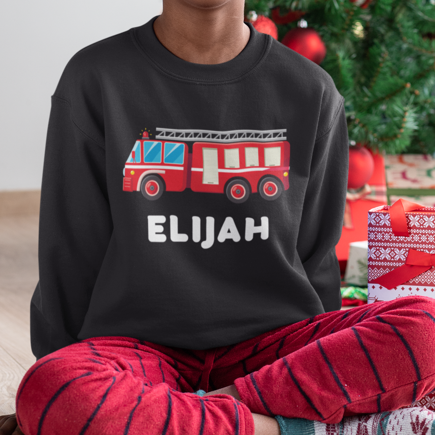 Boy wearing a black sweatshirt with a red fire truck design and 'Elijah' text, sitting in front of a Christmas tree and presents.