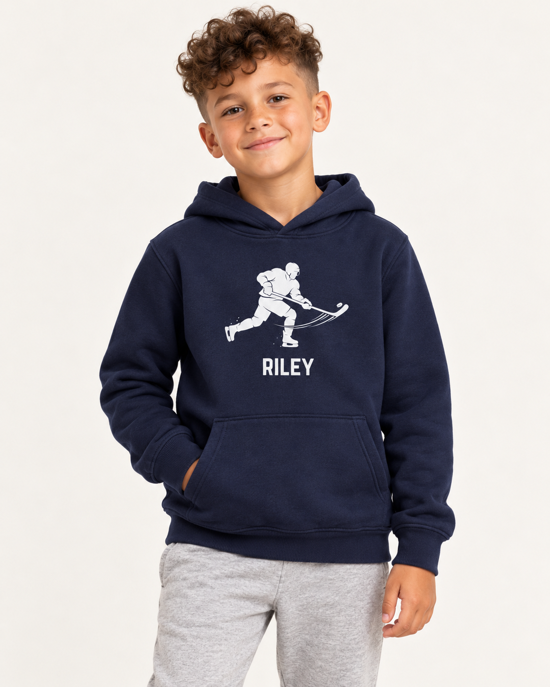 A young boy wearing a navy blue personalised hoodie with a white hockey player graphic and the name 'RILEY' printed on the front, paired with light grey joggers.