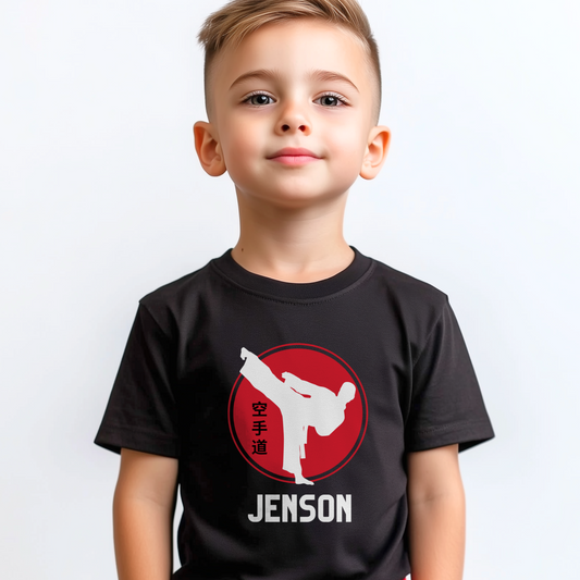 Young boy wearing a black personalised karate T-shirt featuring a white martial artist silhouette performing a high kick inside a red circle with Japanese kanji text, and the custom name “Jenson” printed underneath.