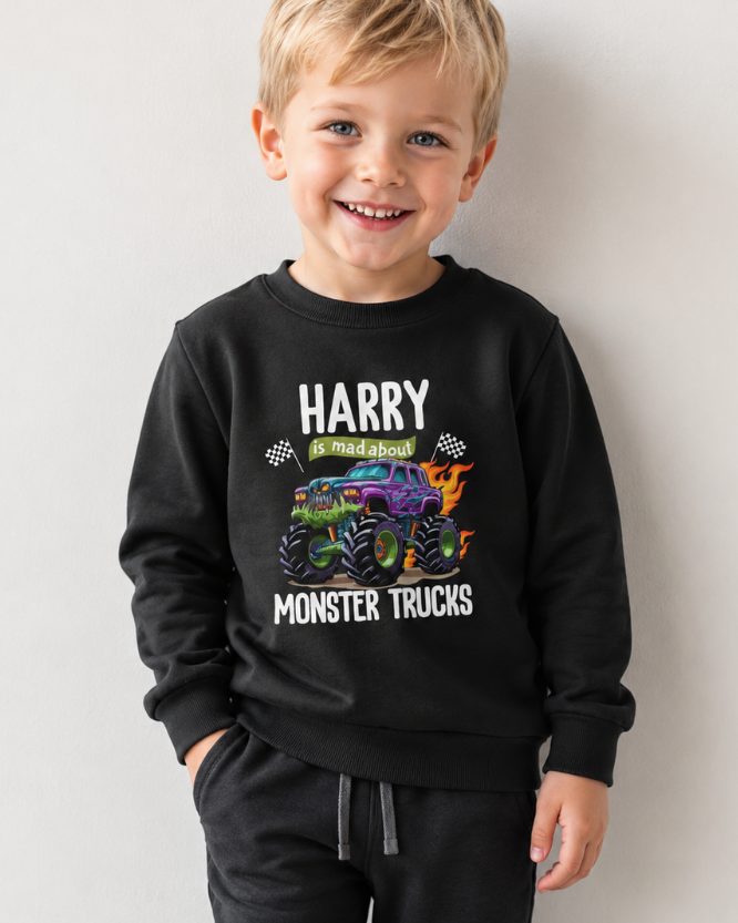 Little boy models a black crewneck sweatshirt featuring a colourful graphic of a purple and green monster truck with flames, accompanied by the text 'HARRY is mad about MONSTER TRUCKS' in white and green.