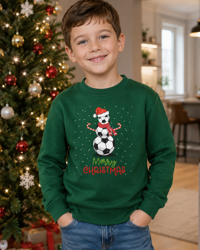 A young boy smiling in front of a decorated Christmas tree, wearing a dark green crewneck sweatshirt with a graphic of a snowman made of soccer balls and the text 'Merry Christmas'.
