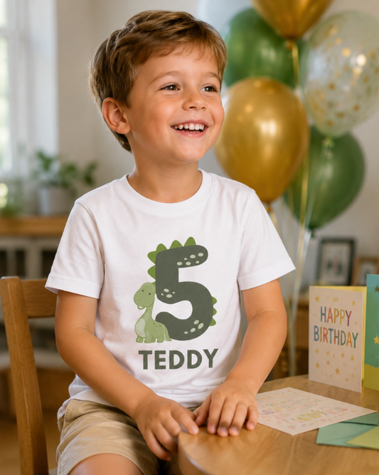 Boy wearing a personalised dinosaur 5th birthday t-shirt with the name Teddy, sitting at a birthday table with green and gold balloons in the background.