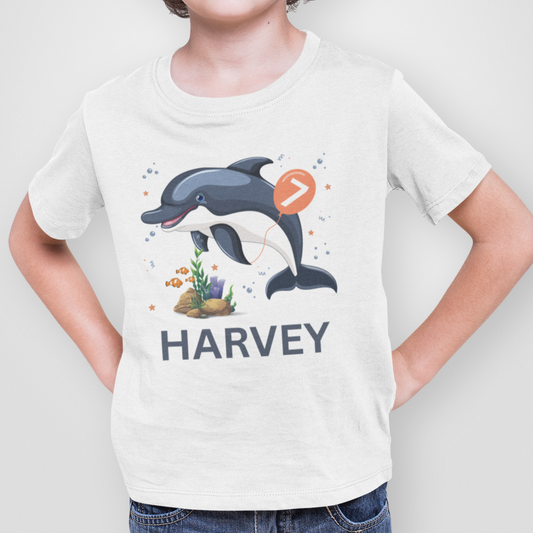 A child wearing a white t-shirt with a cartoon dolphin graphic holding an orange balloon with the number seven, fishes and coral graphic. The name "HARVEY" is printed below.