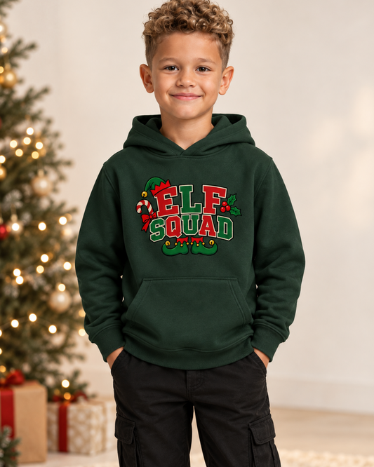 A young boy wearing a dark green pullover hoodie with a colourful 'ELF SQUAD' graphic on the chest, standing in front of a decorated Christmas tree and wrapped gifts.