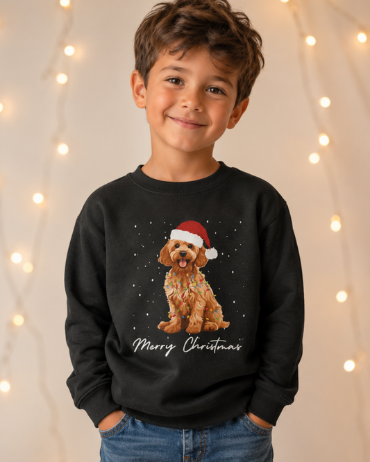 A young boy wearing a black crewneck sweatshirt with a graphic of a golden cockapoo wearing a red Santa hat and the text 'Merry Christmas' underneath, set against a warm background with hanging string lights.
