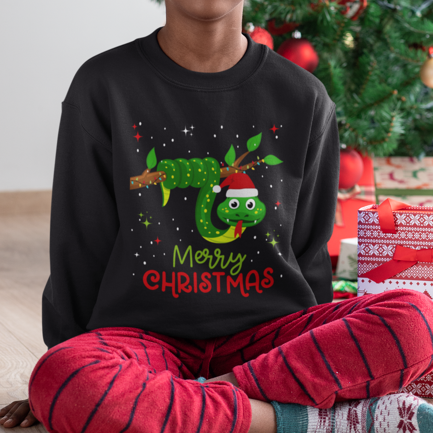 Young boy wearing a Christmas-themed sweatshirt with a snake design and 'Merry Christmas' text, sitting in front of a decorated Christmas tree and presents.