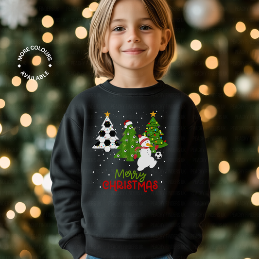 Boy wearing a black Christmas-themed sweatshirt with trees, snowman kicking a soccer ball design, boy is standing in front of a decorated Christmas tree.