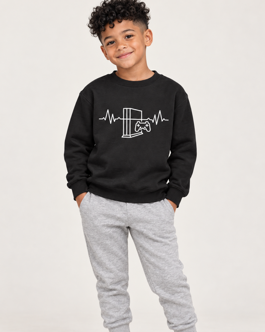 A young boy models a black crewneck sweatshirt with a white graphic of a gaming console and controller with a heartbeat line, paired with heather grey joggers.