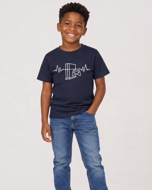 A young boy smiling and wearing a navy blue crewneck t-shirt with a white video game console and heartbeat graphic, paired with blue jeans, against a plain light gray background.