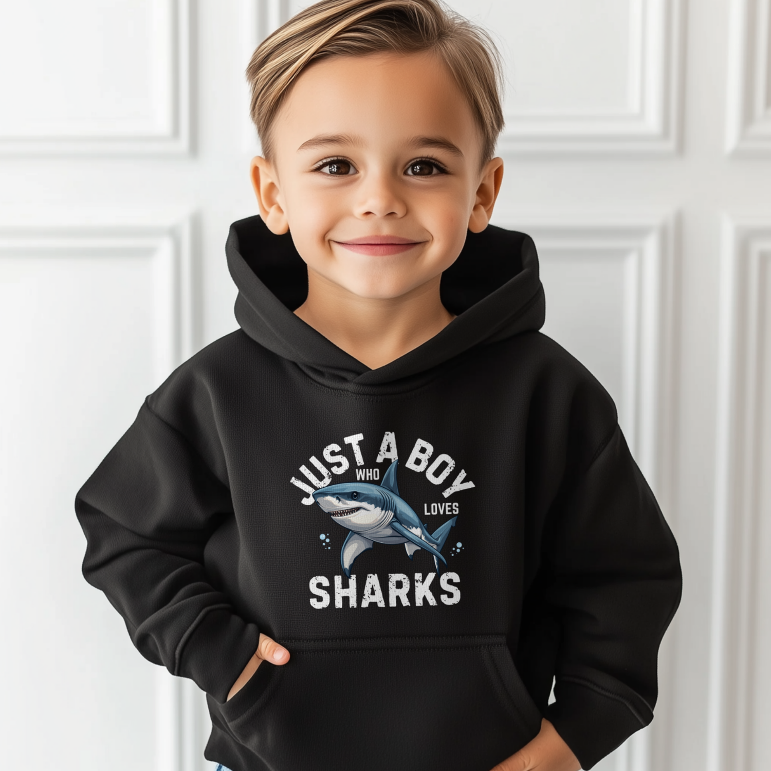 Little boy wearing a black hoodie with a shark graphic and text, standing against a white panelled wall.
